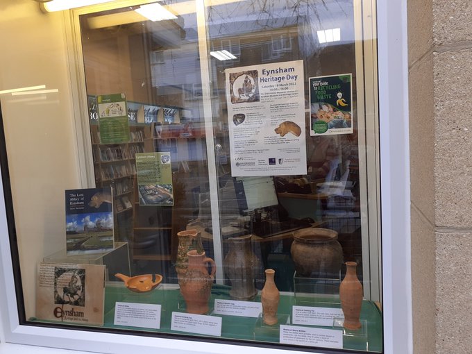 A window display of ancient Eynsham artefacts, with a poster advertising Eynsham Heritage Day in March 2022