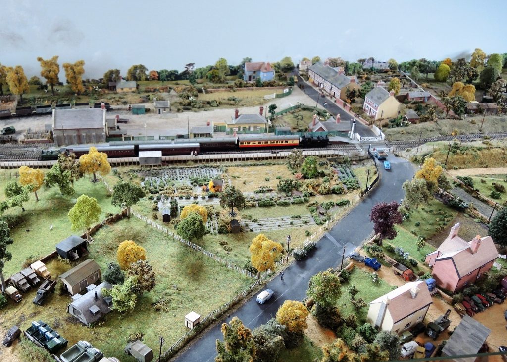 Model of Eynsham Station in the 1950s, created by David Thomas. View from above of a passenger train at the station, with houses, allotments, fields, trees in the vicinity.