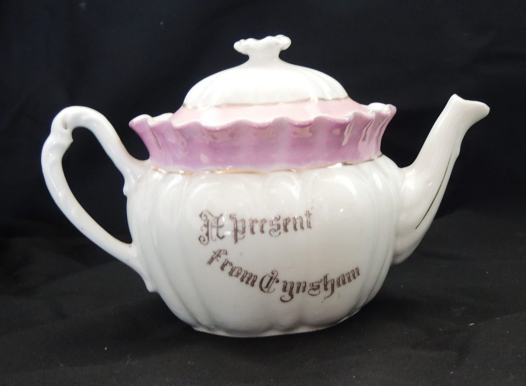A white and pink fluted teapot with gold lettering that says 'A present from Eynsham'.