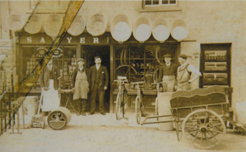 Monochrome photo of the front of Sawyers Shop on Newland Street, Eynsham. A group of people are standing outside the shop, alongside an empty two-wheeled cart.
