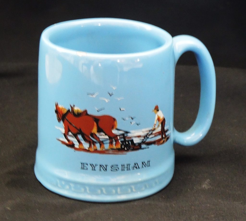 A pale blue mug dating from the 1960s, with an image of a horse-drawn plough and a man, with the word Eynsham underneath,